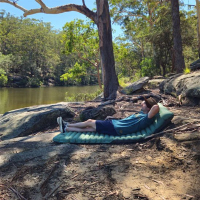 Homme couché sur un matelas gonflable 1 personne auto gonflant, sous un arbre près de l'eau.