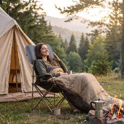 Devant sa tente une femme se repose avec la tête un oreiller gonflable ergonomique compact gris, devant sa tente, près du feu.